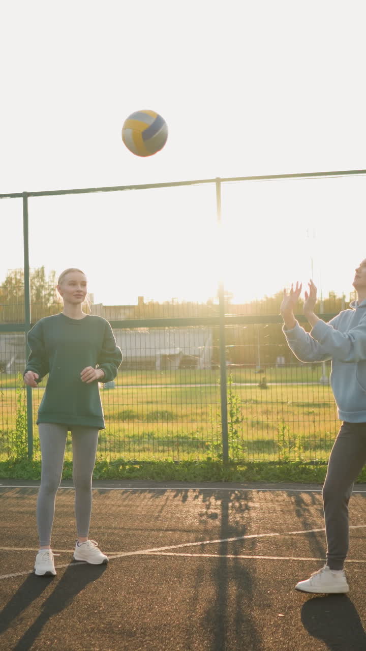 초보자 배구 선수들이 야외에서 연습을 하고 있으며, 배경에는 축구장이 있고, 한 선수가 공을 때리기 위해 준비하는 동안 따뜻한 빛이 긴 그림자를 던집니다.