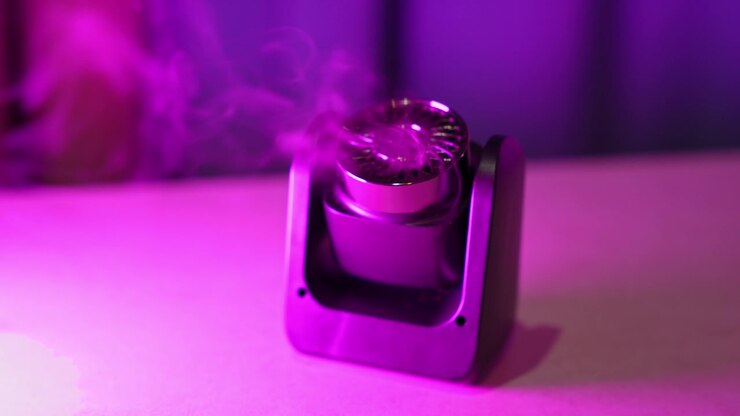 Nærbilde av en liten svart elektronisk aromadiffusor som sender ut en svak stripe med damp. Innendørs studiomiljø opplyst av levende rosa og lilla neonlys mot en mørk, uskarp bakgrunn