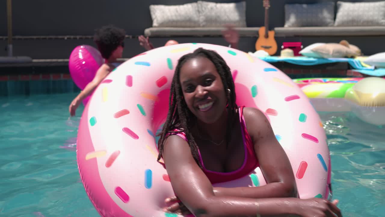 African friends chatting in backyard pool, woman putting on sunglasses, leaning through donut float
