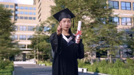 asian woman student graduates in cap and gown with diploma shaking head and disapproving with no index finger sign in front of a magnificent university building