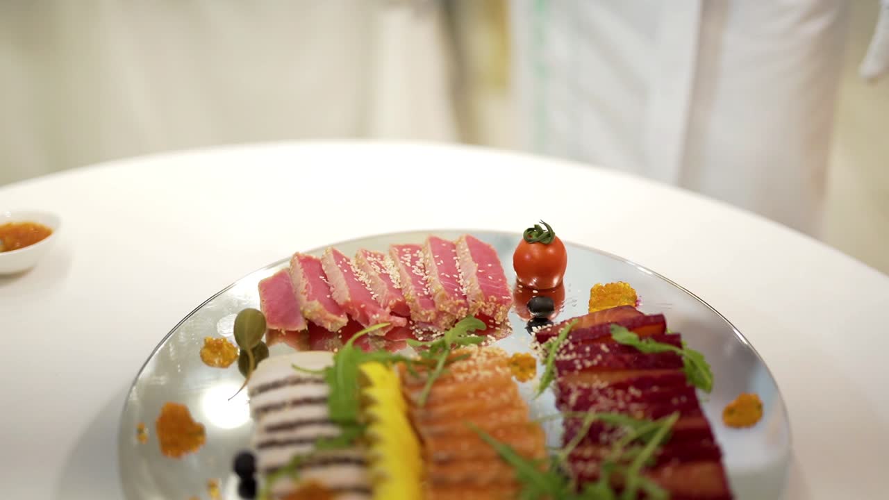 Chef's hand adds cherry tomato to a tasty appetizer of meat on a glass plate on a white table. A cook in white clothes decorates slicing snack for the banquet.