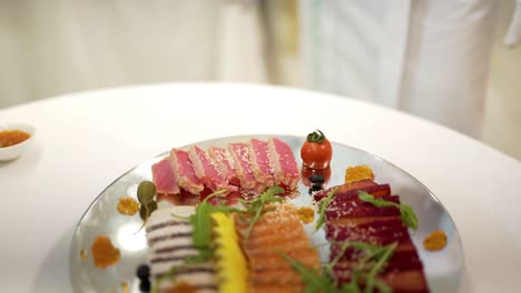 chef's hand adds cherry tomato to a tasty appetizer of meat on a glass plate on a white table. a cook in white clothes decorates slicing snack for the banquet.