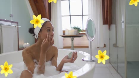 Animation-of-yellow-flowers-over-biracial-woman-relaxing-in-bath-moisturising-face-at-home