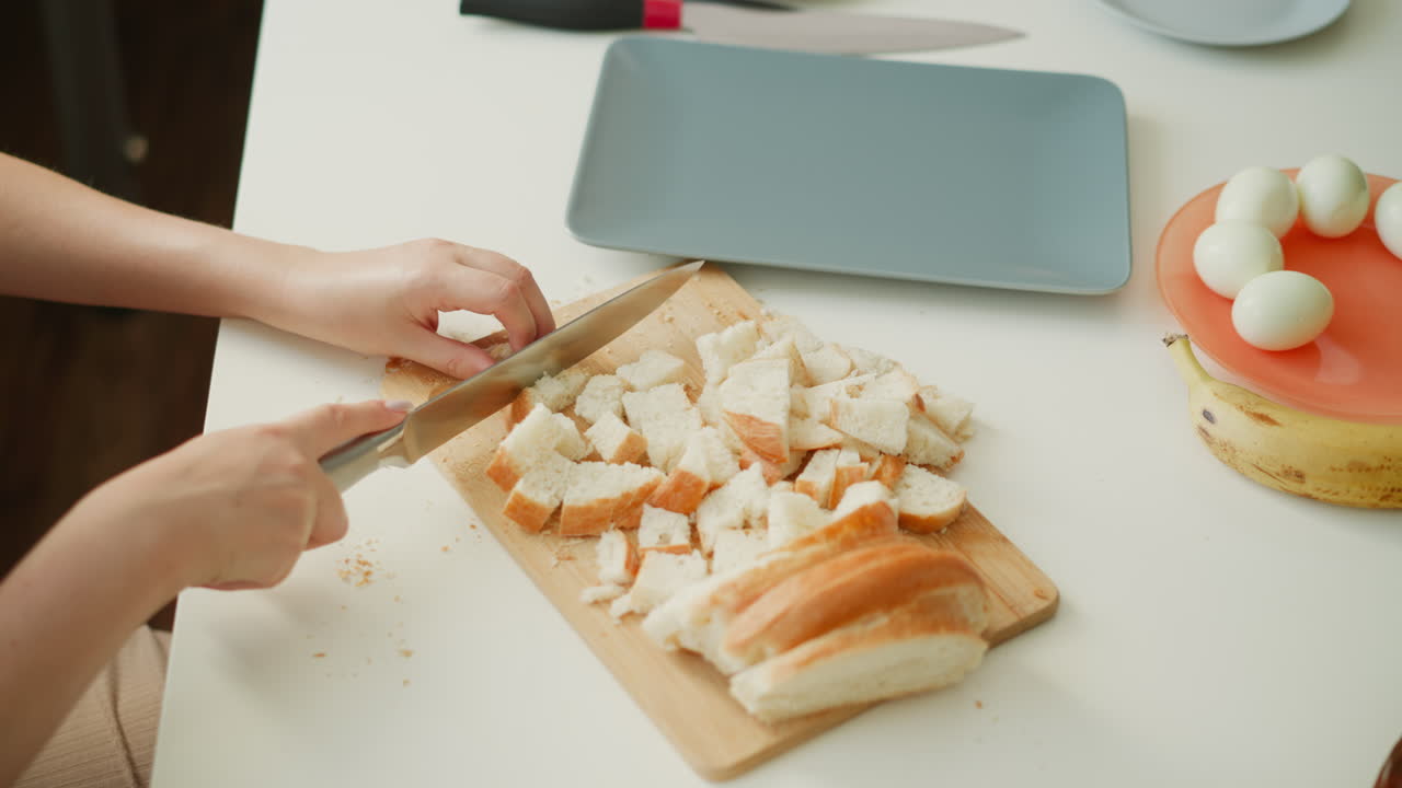High angle view of culinary artist chopping bread slices into precise small cubes on wooden board over white table under soft window light, crumbs scattered, capturing cooking preparation process