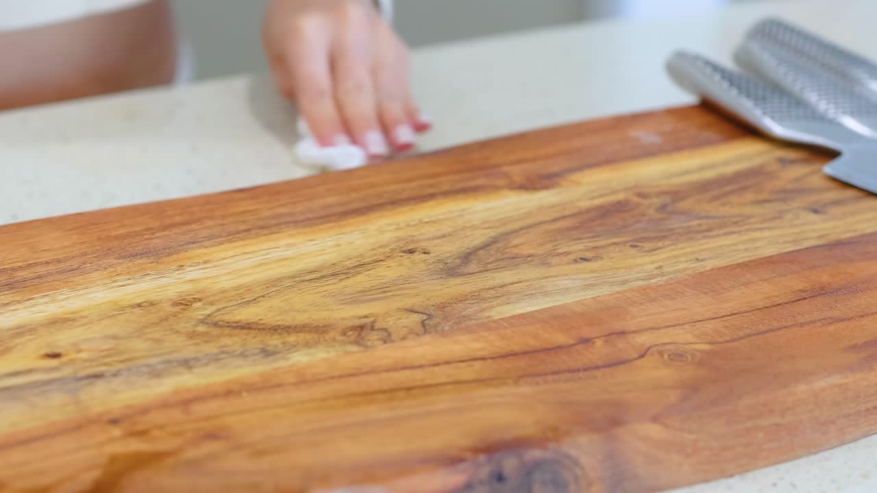 A person wipes a wooden cutting board with a cloth in a well-lit kitchen, surrounded by fresh tomatoes and knives