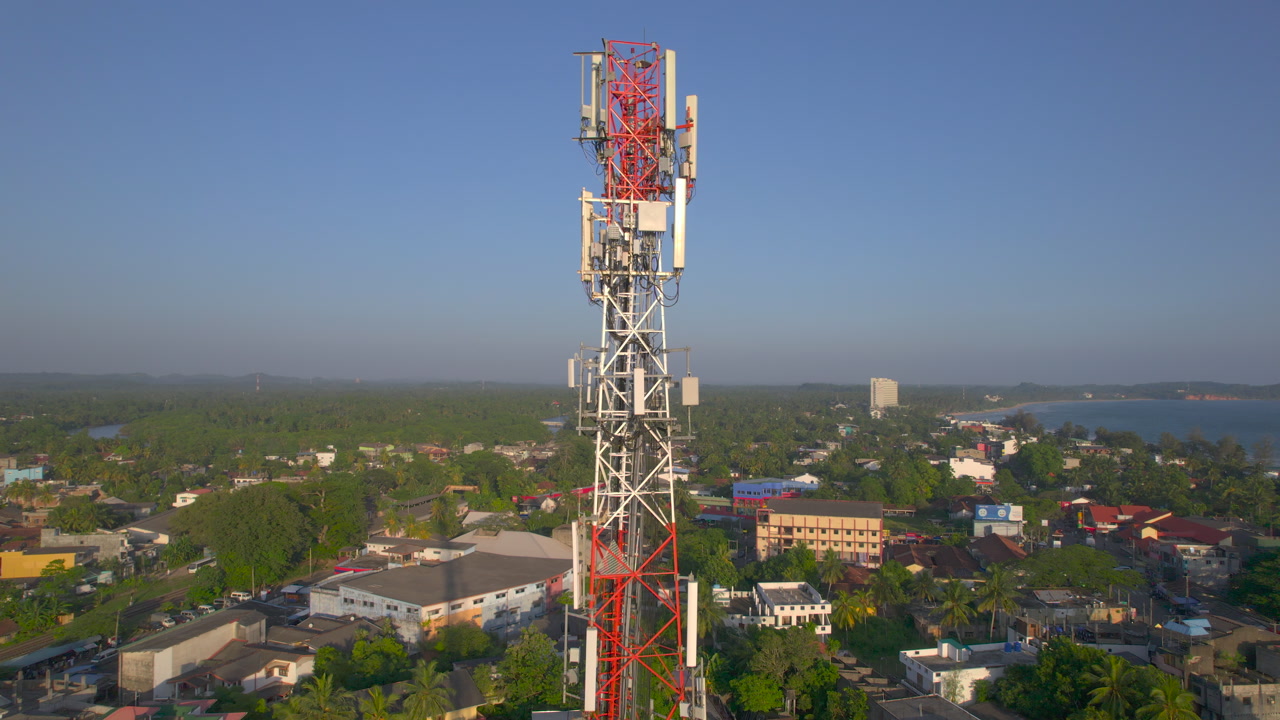 Aerial video of telecom cell tower rising above tropical coastal town with palm trees seaside buildings and calm bay communication infrastructure and shoreline landscape in daylight close view