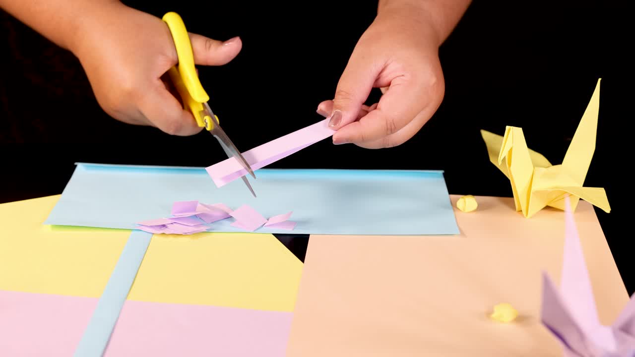 Person uses scissors to cut colored paper strips for origami on pastel workspace, overhead view