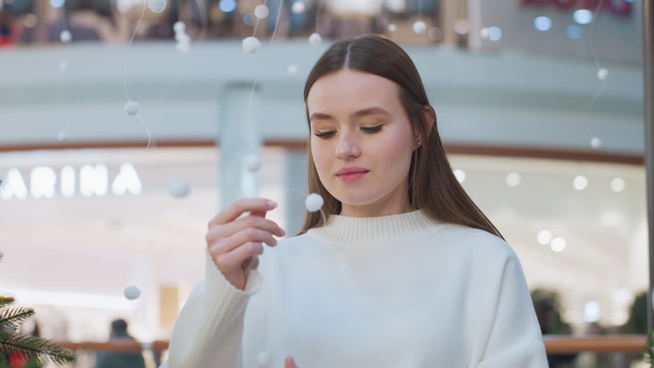 Young lady in elegant white sweater stands in festively decorated shopping mall, gently holding delicate hanging ornament, surrounded by soft lights, she admires the festive ambiance