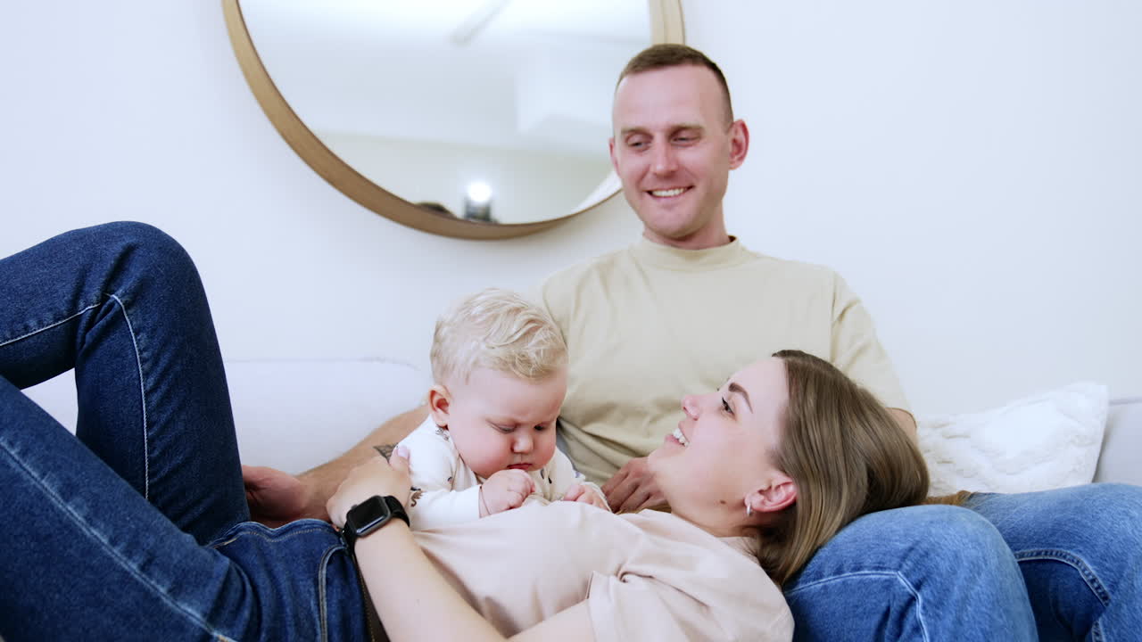 Happy Caucasian parents having rest together with their baby. Little kid sits by his mom lying on husband's laps. Proud dad caresses son's head.