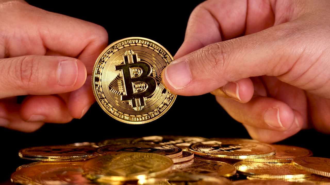 Hand passes gold Bitcoin coin to another hand above pile, dramatic lighting, close-up view