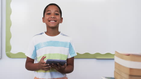 Un-Niño-Birracial-Con-Una-Camisa-A-Rayas-Sostiene-Una-Tableta-En-La-Escuela,-Sonriendo-En-Un-Salón-De-Clases