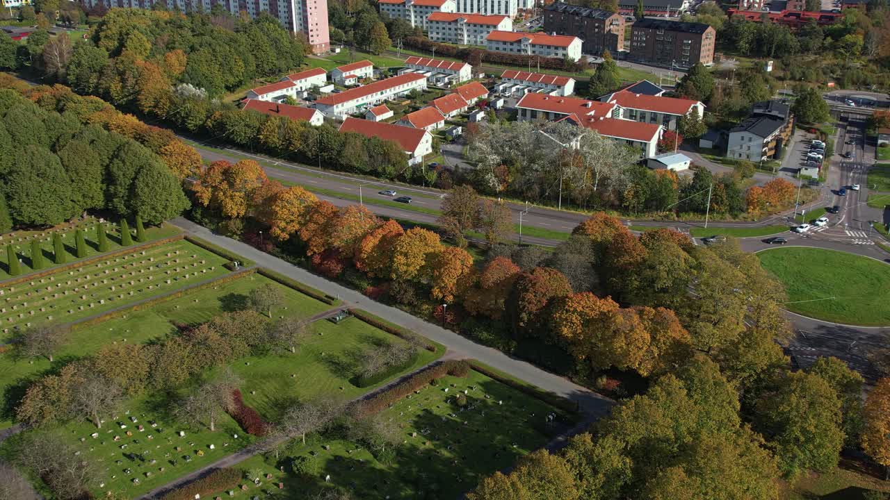 Slow aerial orbit of Kviberg cemetery grounds, fall trees, and nearby red roofed residential area in Gothenburg, Sweden