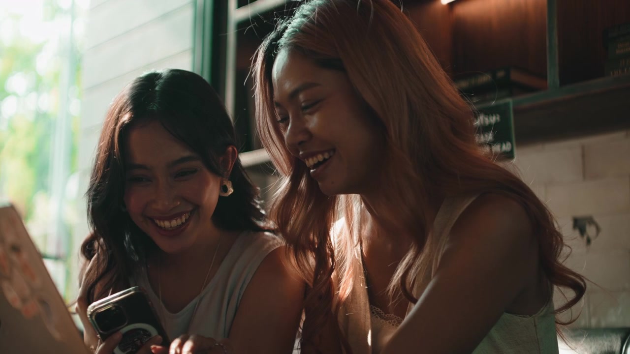 カフェで笑いながらスマートフォンを使っている若い女性二人。
