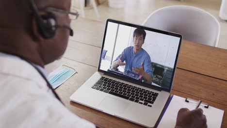 african american male doctor wearing phone headset taking notes while having a video call on laptop