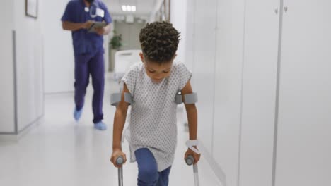 african american male doctor walking with child patient using crutches at hospital