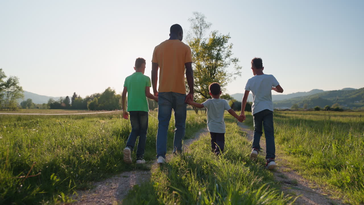 Vista traseira de um pai afro-americano e seus três filhos caminhando juntos de mãos dadas ao longo de um