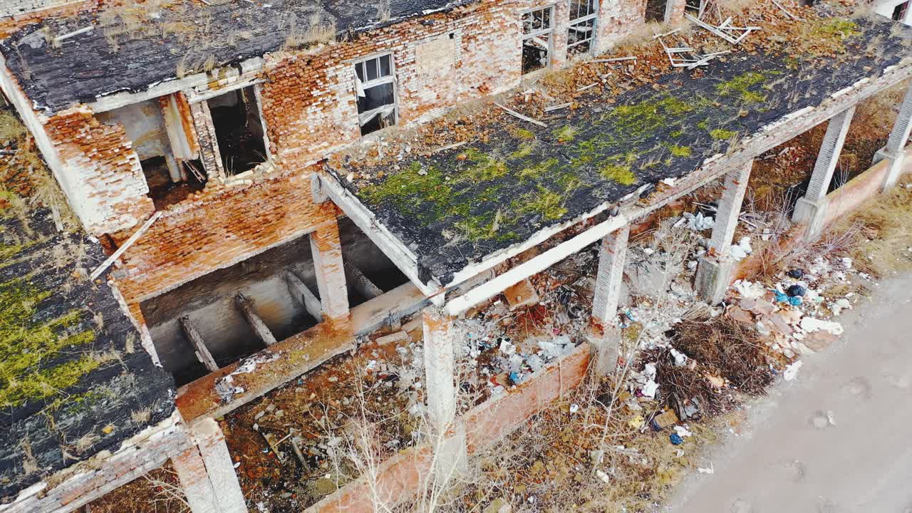 Ruined brick buildings with collapsed roofs. Broken dirt road with heaps of garbage. Moss and grass on the ruins. Aerial view