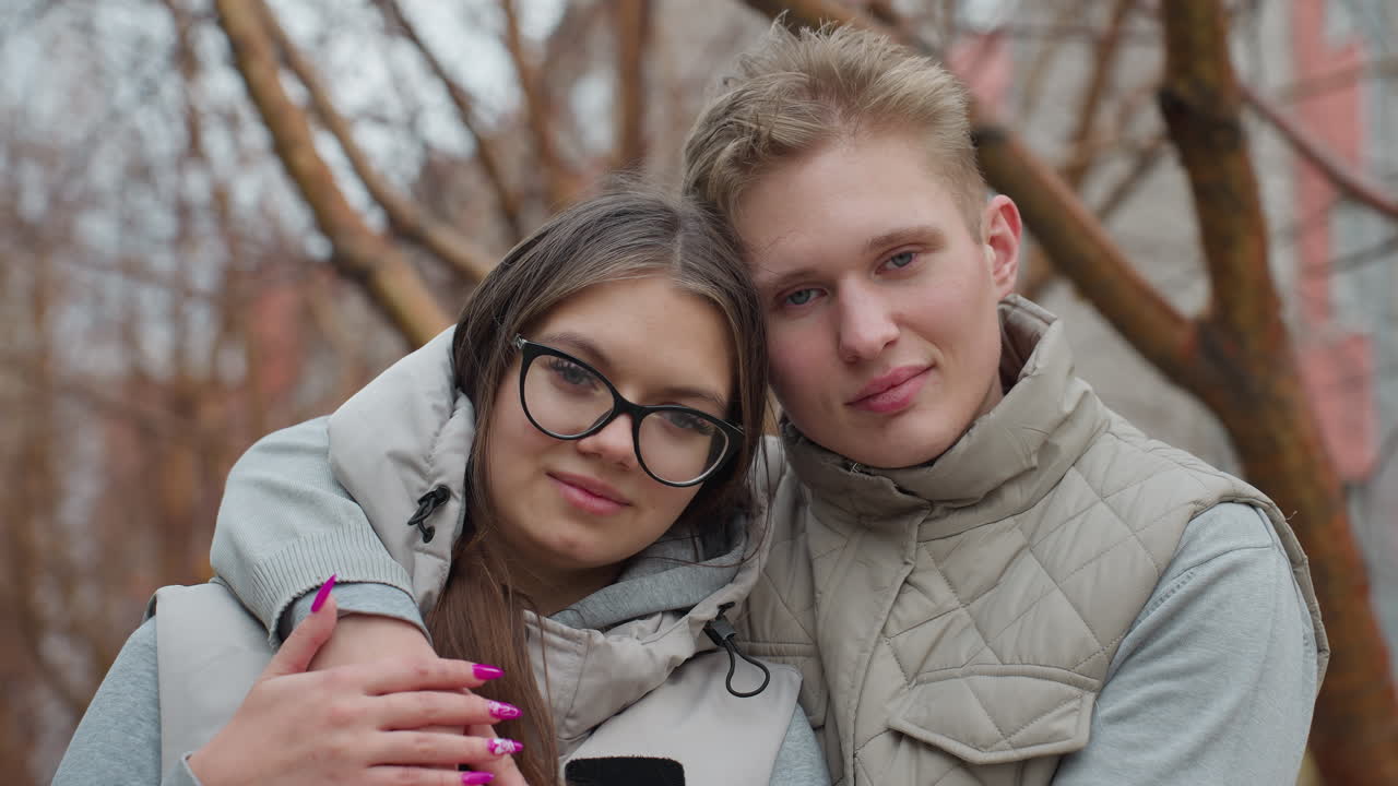 Beautiful moment of lovely couple holding each other warmly in cozy outdoor setting, standing close together with gentle expressions as trees without leaves sway behind them