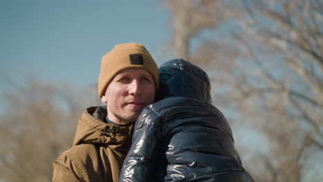 a front view of a father carrying his son while walking, the father is dressed in a brown jacket and beanie, while the child is snug in a black puffy jacket, with blurred the trees and sky