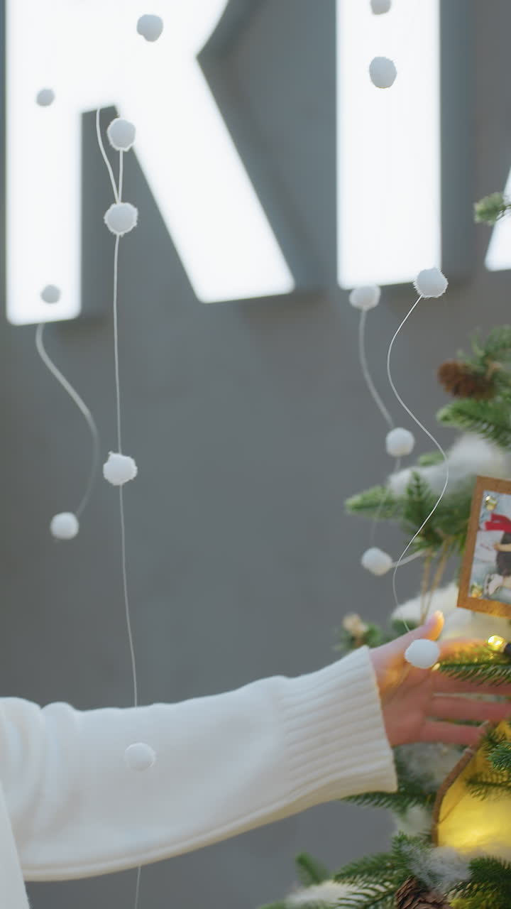 Serene woman in white sweater touches a picture ornament on a Christmas tree, admiring the illuminated inscription, surrounded by festive lights and decor in a mall setting