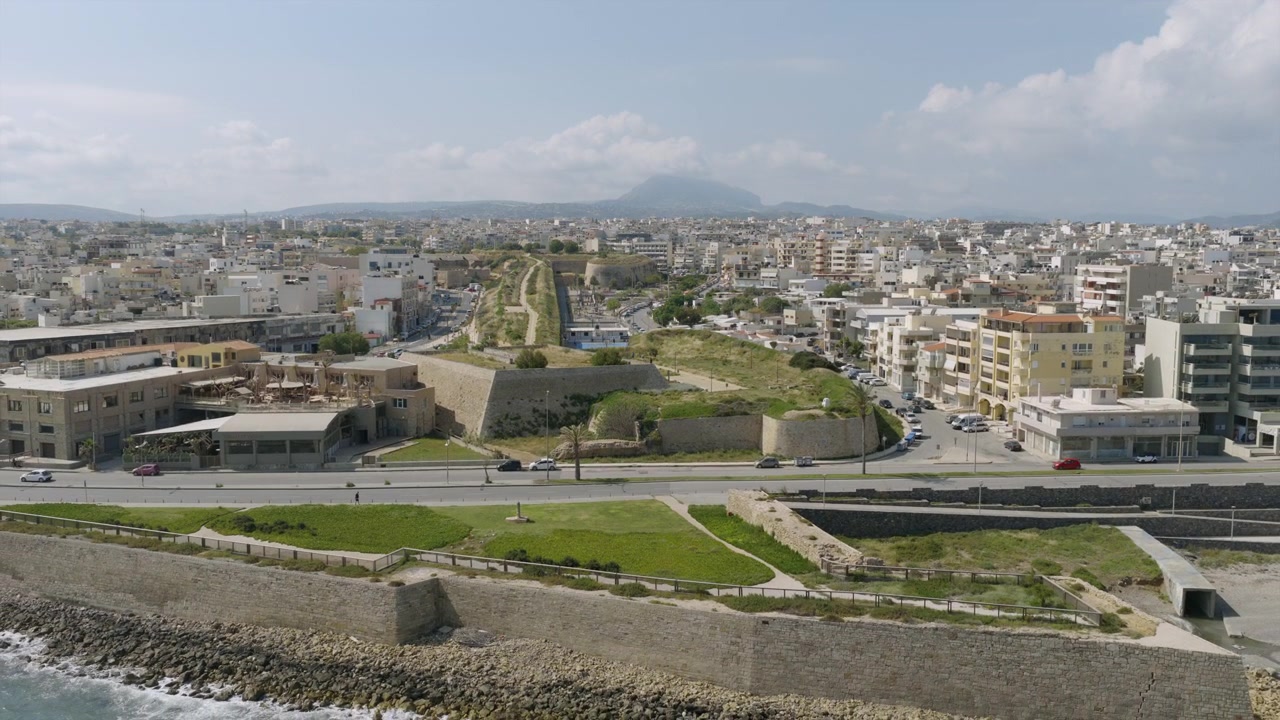 Vista aerea dei bastioni veneziani ricoperti d'erba di Heraklion, Creta, con una strada trafficata sottostante e gli edifici della città che si estendono verso montagne sfocate.