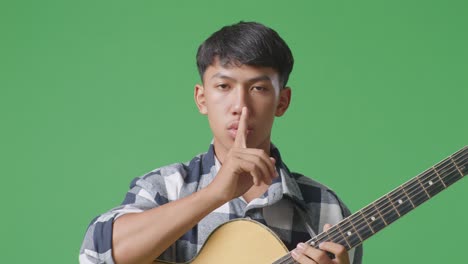 primer plano de un adolescente asiático con una guitarra mostrando un gesto de silencio mientras está de pie en el fondo de la pantalla verde