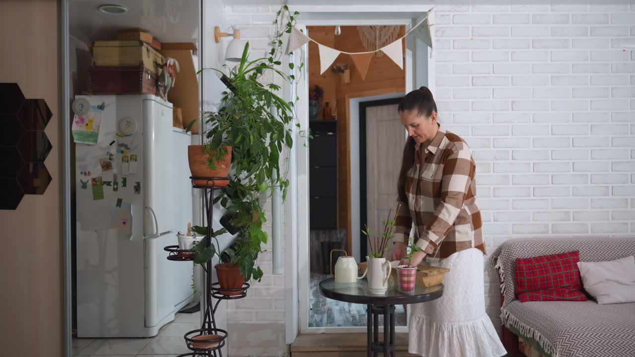 Gardener barefoot walks into cozy modern room carrying wooden tray places tray on round table next to potted plants and sofa sheer curtains framing snowy landscape through glass door