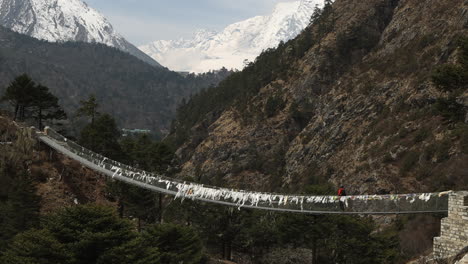 person walking slowly across suspension bridge in nepal with mountains in the background.