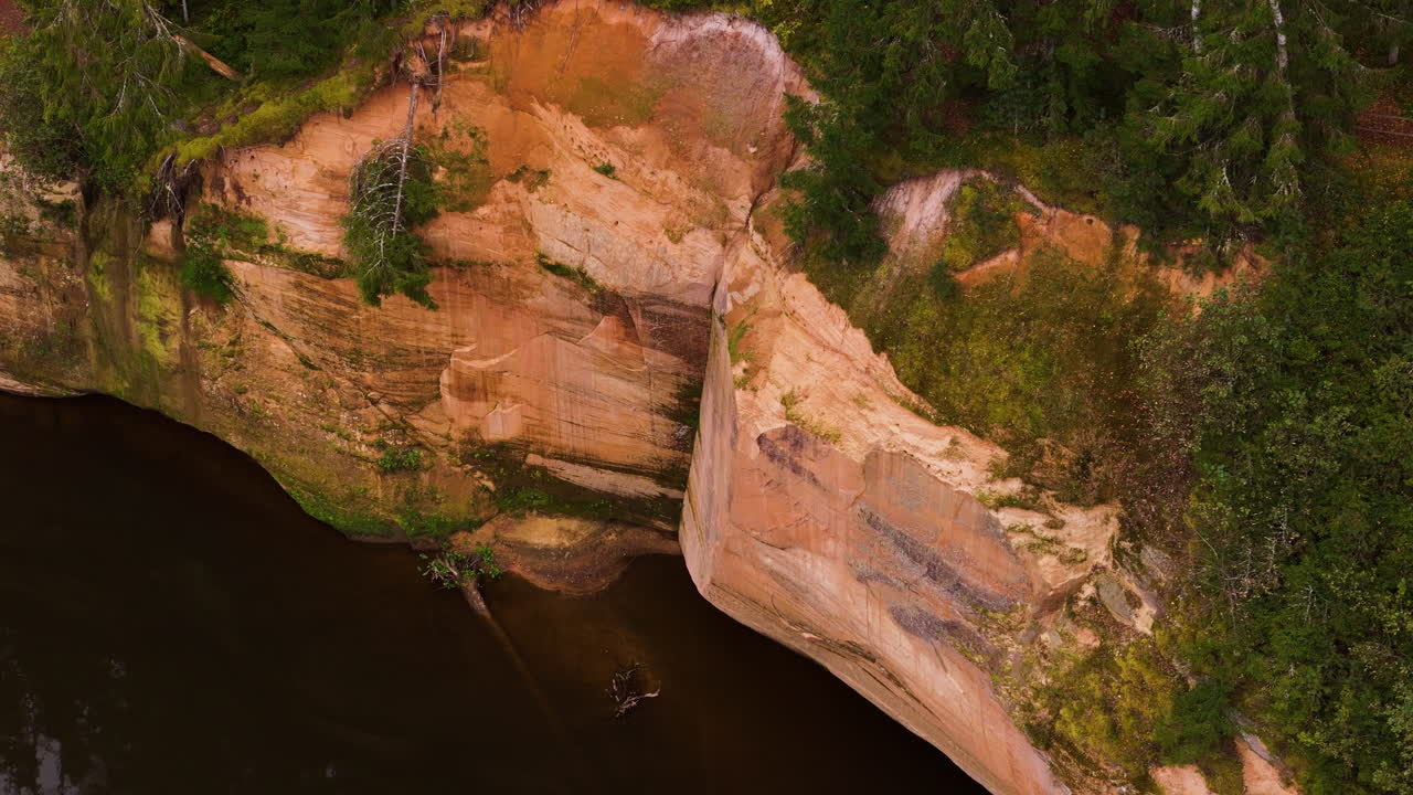 Aerial footage of vertical Erglu sandstone cliffs above dark river water in Latvia.