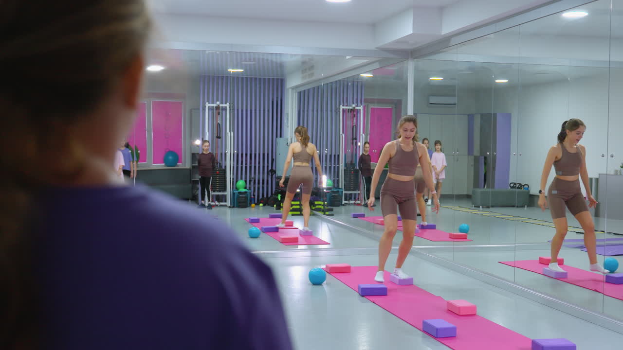 보라색 옷을 입은 여성이 한 명의 강사가 한 다리에 균형을 잡는 운동을 시연하는 것을 지켜보고 있으며, 거울은 배경, 다른 참가자 및 체조 장비를 반영합니다.