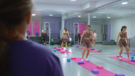 vista trasera de cerca de una mujer de púrpura viendo a un instructor demostrar un ejercicio de equilibrio en una pierna, con un espejo que refleja el fondo, otros participantes y equipos de gimnasio