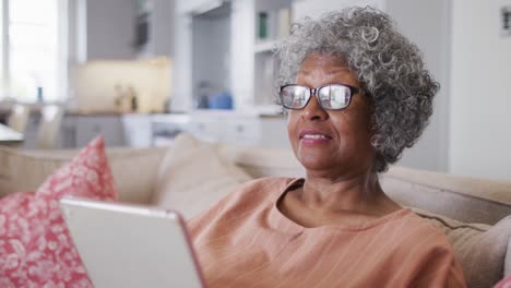 Ältere-Afroamerikanische-Frau,-Die-Einen-Videochat-Auf-Einem-Digitalen-Tablet-Führt,-Während-Sie-Auf-Der-Couch-Bei-Ho-Sitzt