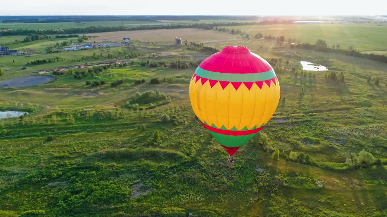 Beautiful aerostat against the evening sunlight. Colorful round hot air balloon flying over the green nature. Airship in the air in rural place at sunset. Aerial view.