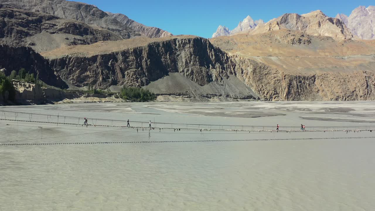 drones aéreos volando hacia atrás revelando las montañas de conos passu en la distancia mientras los turistas cruzan con cautela el famoso puente hussaini en hunza pakistán mientras un río gris de ritmo rápido fluye debajo