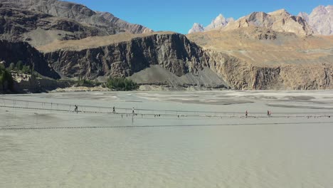 aerial drone flying backwards revealing the passu cones mountains in the distance as tourists cautiously cross the famous hussaini bridge in hunza pakistan as a fast paced gray river flows below