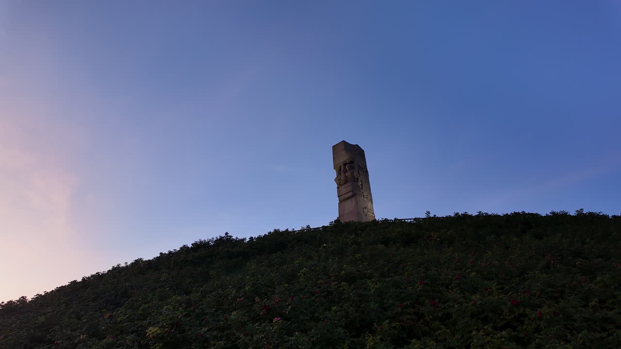 A wide, low-angle shot of the Westerplatte monument standing atop a lush green hill against a serene dawn sky in Gdańsk, Poland. A historic WWII landmark