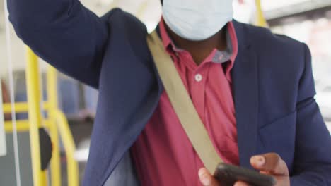 Midsection-of-african-american-man-in-city-wearing-face,-using-smartphone-in-bus