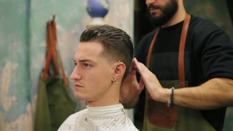 Vista-Lateral-De-Un-Joven-Apuesto-Hombre-Caucásico-Con-Un-Piercing-En-La-Oreja-Vistiendo-Su-Cabello-Y-Peinándolo-Por-Un-Barbero-Barbudo-En-Un