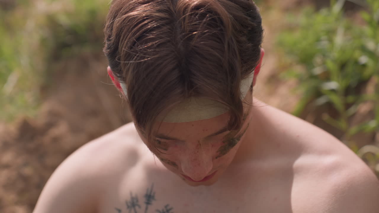 Fierce young warrior with intense glare and muddy streaks on cheeks gazes forward, wrapped headband across forehead, standing shirtless under sunlight, embodying primal strength, and raw emotion