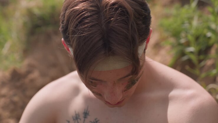 Fierce young warrior with intense glare and muddy streaks on cheeks gazes forward, wrapped headband across forehead, standing shirtless under sunlight, embodying primal strength, and raw emotion