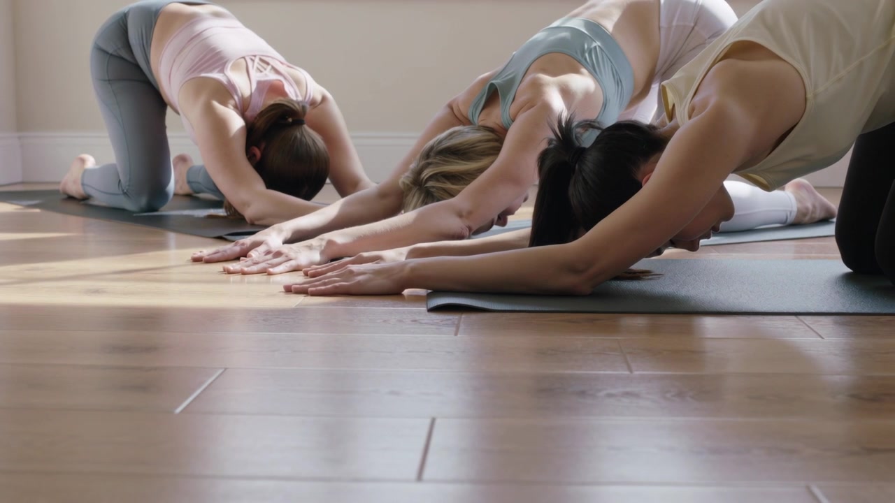 Espirando, tre donne si sistemano nella posizione del bambino in uno studio, su tappetini, durante una lezione in abbigliamento sportivo.