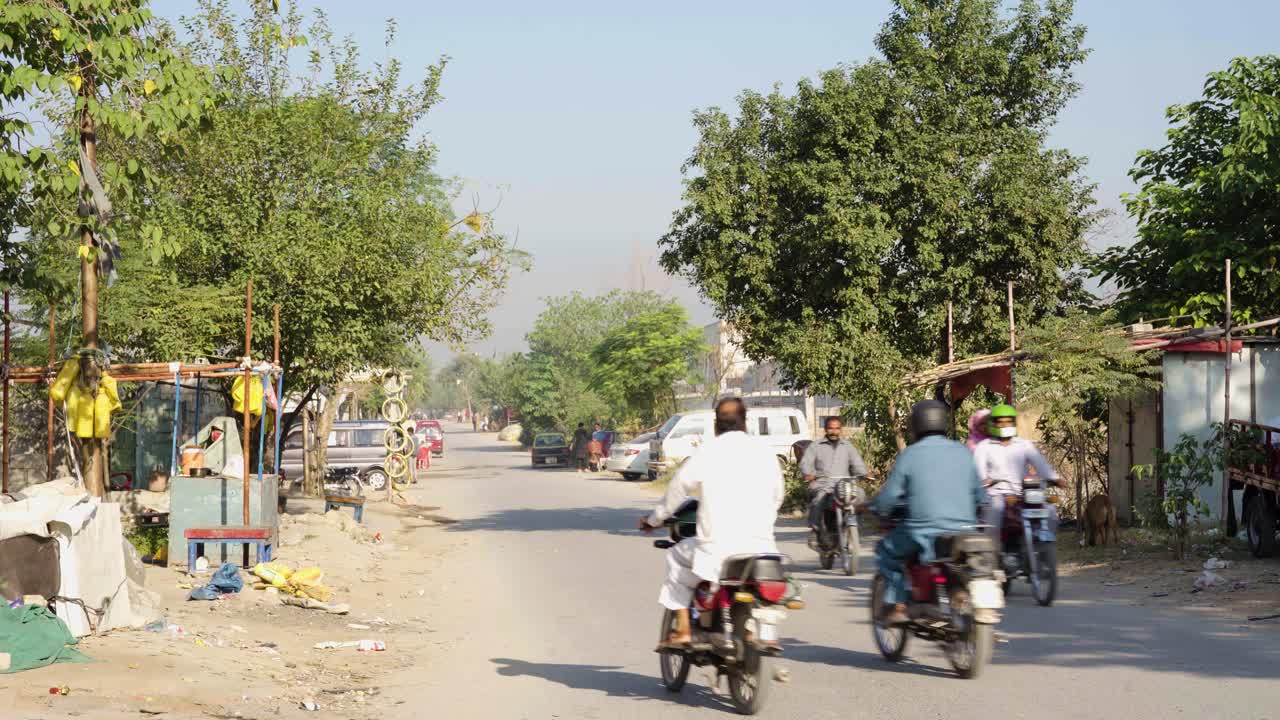 A rural street scene in a neighborhood of a Pakistani city, showcasing animal stores and pedestrians.