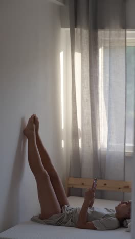 Woman relaxing on bed with legs up and using phone