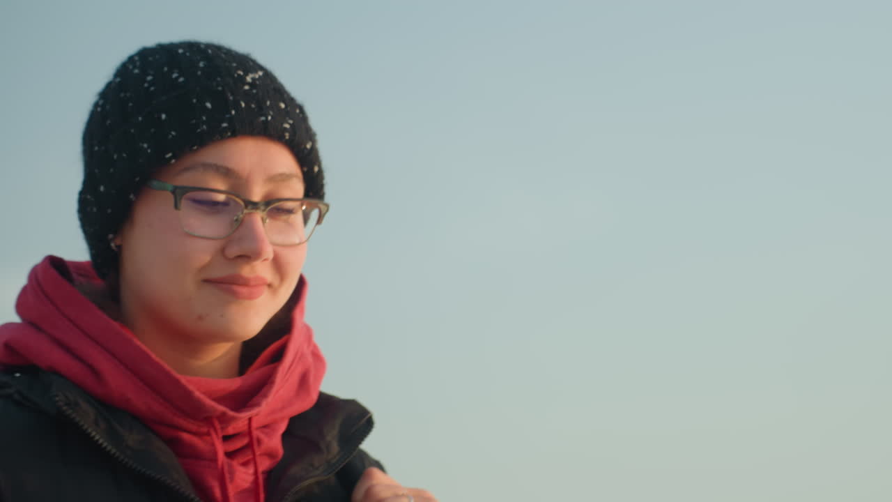 Junge Frau blickt am Strand bei Sonnenaufgang, mit Mütze und Brille, roter Kapuzenpullover, ruhiges, nachdenkliches Porträt mit sanftem Himmel, fernen Wellen, kalter Luft und leichten Schneeflocken auf der Mütze, eine studentische Reisende, die tief durchatmet.