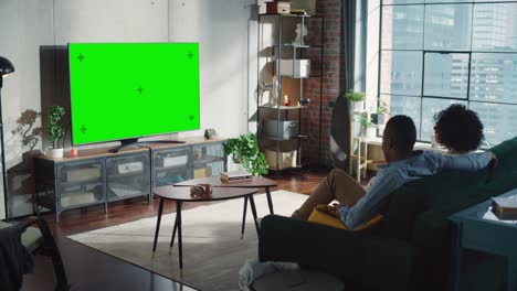 black couple sitting at home in the living room and watching green chroma key screen television, relaxing on a couch. weekend entertainment concept