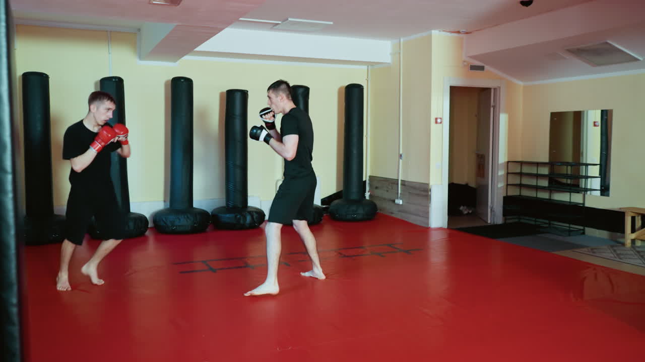 Wrestlers sparring inside gym, wearing gloves and black outfits, practicing combat sport moves with focused stance and movement on red mat floor surrounded by punching bags during intense martial training