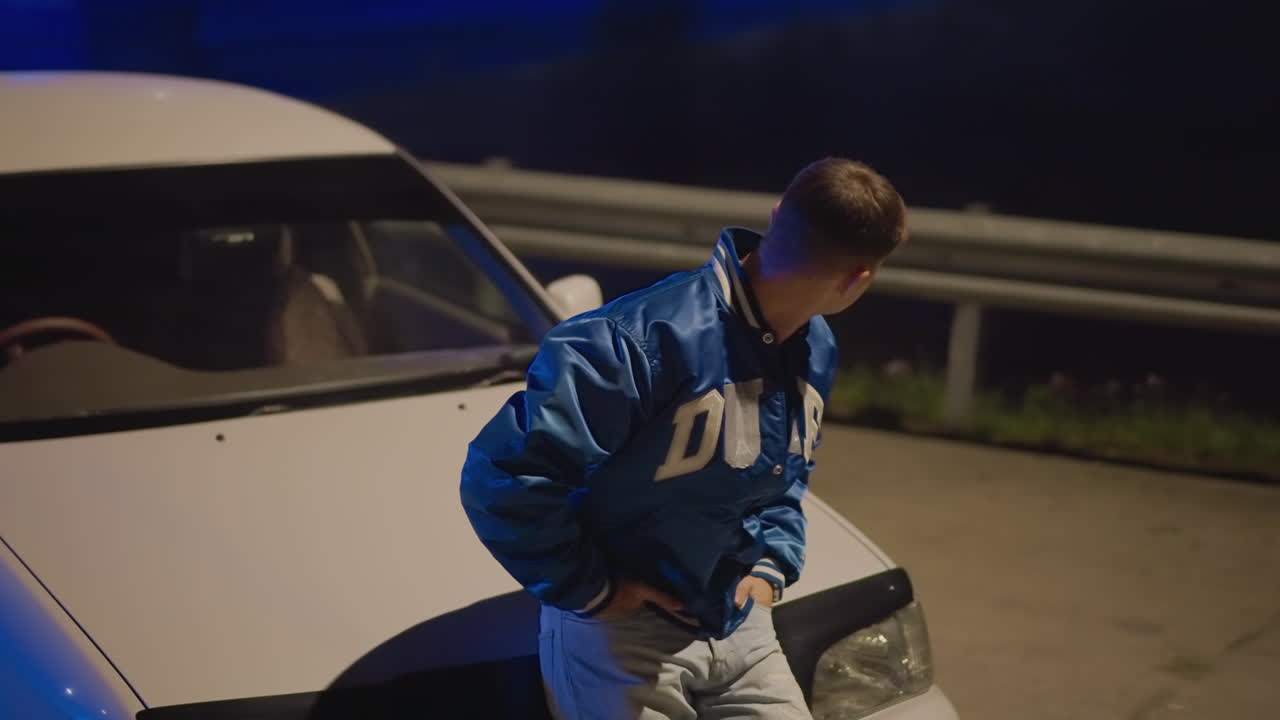 Night roadside portrait white driver leaning on car, pensive young man in blue jacket under streetlight, empty highway and guardrail in background, headlights and cool blue tint, cinematic moody