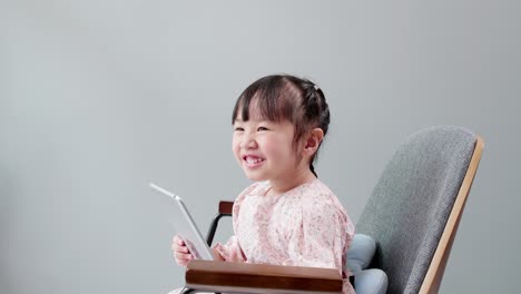 Niño-Asiático-Con-Flecos-En-El-Cabello-Sentado-En-Una-Silla-Riéndose-Mientras-Usa-Una-Tableta-Digital