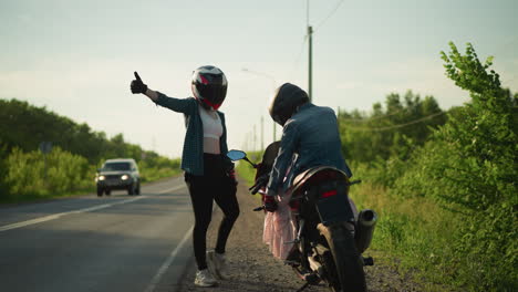 two women by the roadside, one seated on a motorcycle playing with her clothes while the other stands and gives a thumbs up, a car approaches in the distance