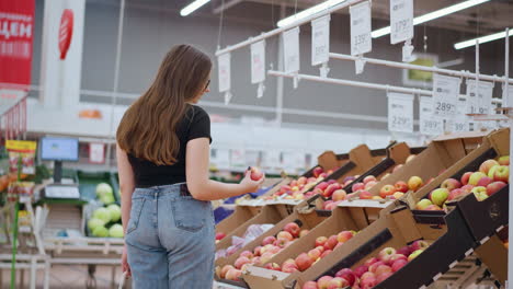 rear view of lady with phone in back pocket picking red apple from display, observing it, and dropping it back, price tags and signs visible in the background of bright store environment